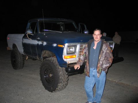1995 Ford Bronco - A man poses next to a modified pickup truck with oversized tires and a two-tone paint job.