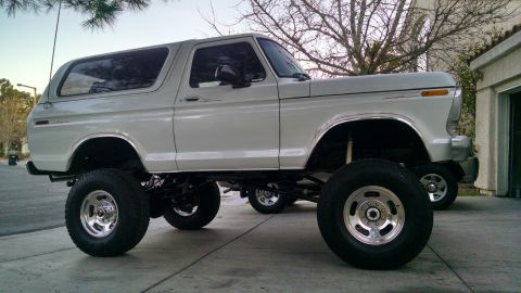 1979 Ford Bronco - A lifted, white 1979 Ford Bronco with large off-road tires is parked on a concrete driveway. The vehicle is viewed from the side, showcasin…