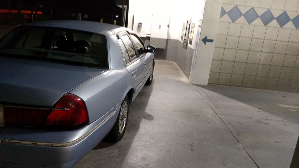 Rear passenger side of a light blue sedan parked in a concrete area with a tiled wall and a blue arrow.