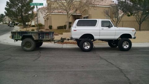 1979 Ford Bronco - A lifted white 1970s Ford Bronco is towing a military-style trailer on a residential street. The Bronco has large aftermarket wheels and ti…