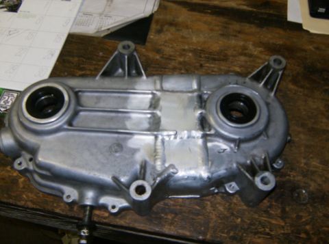 2000 Ford E350 - A silver metal engine part, possibly a transmission or timing cover, is shown on a wooden surface. The part has two circular openings with…
