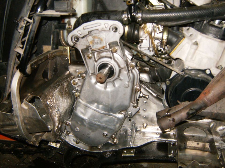 Close-up of engine and transmission parts, possibly from a snowmobile or similar vehicle. Shows metal casing, hoses, and some wiring