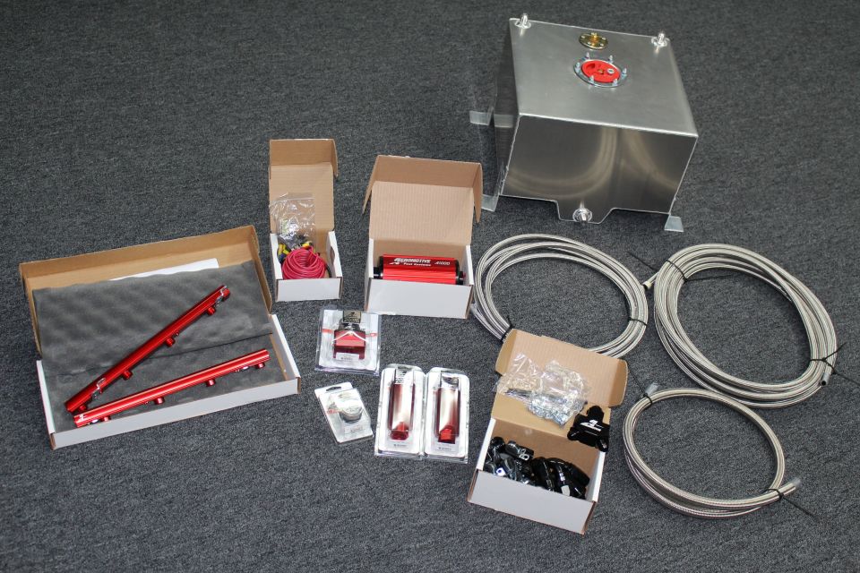 Aeromotive fuel system kit laid out on gray floor: aluminum fuel cell, red rails, regulator, gauges, and braided line coils