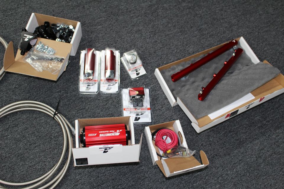 Boxed fuel system parts on gray carpet: black fittings, red gauges, red A1000 pump, red shocks, and coiled wire