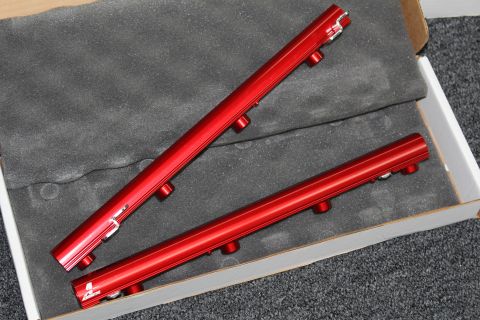2003 Ford Crown Victoria - Two red anodized aluminum fuel rails laid diagonally on gray foam packaging inside a cardboard box.
