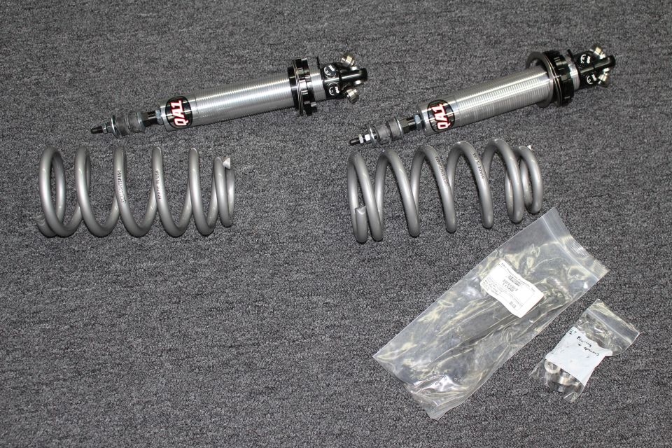 Pair of polished coilover shocks, two gray coil springs, and bagged hardware on carpet