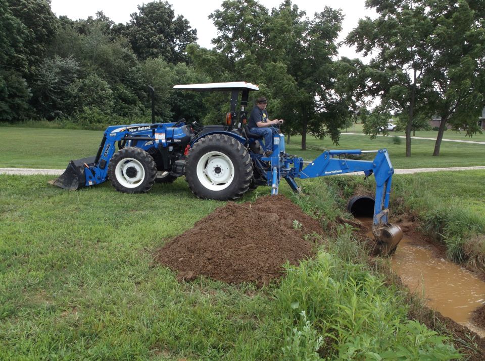 Blue New Holland tractor with front loader and backhoe attachment digging a ditch next to a muddy stream