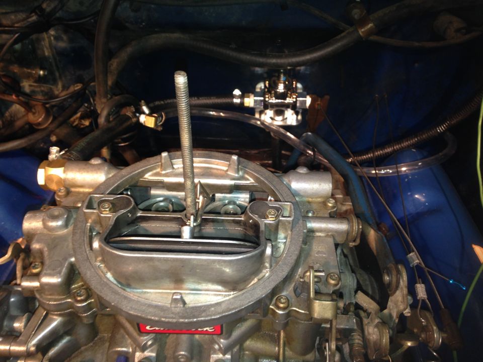 Close-up of a carburetor with fuel lines connected. A metal rod extends upwards from the center