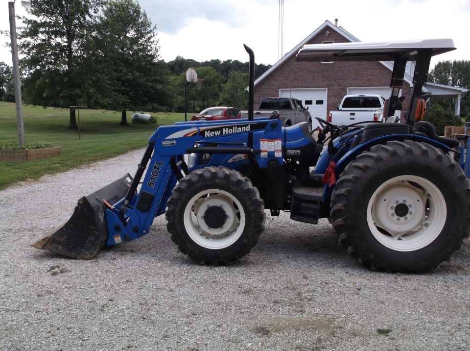 Blue New Holland tractor with a front loader parked on gravel. Two pickup trucks and a car are visible in the background