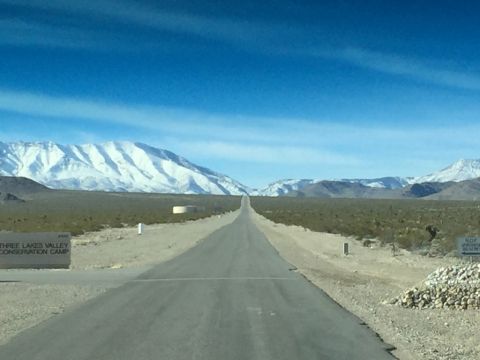 1979 Ford Bronco - A long, straight road cuts through a desert landscape towards distant snow-capped mountains, with a sign for Three Lakes Valley Conservatio…