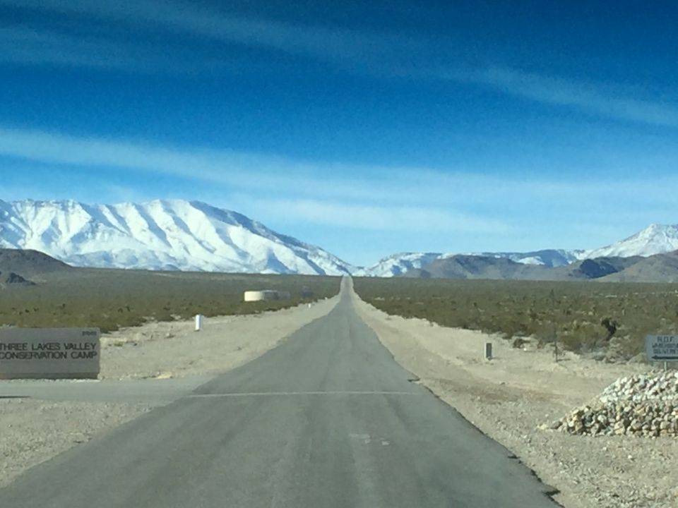 Desert road leading to snow-capped mountains, sign for Three Lakes Valley Conservation Camp