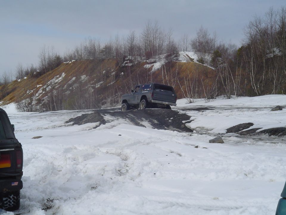 Two SUVs driving on a snowy, hilly terrain with bare trees in the background