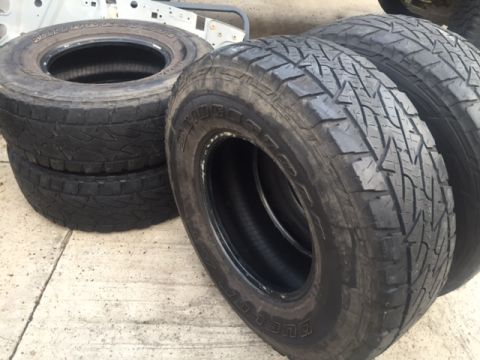 1986 Ford Bronco - Four Bridgestone Dueler tires are stacked on a concrete surface, with a vehicle frame visible behind them.