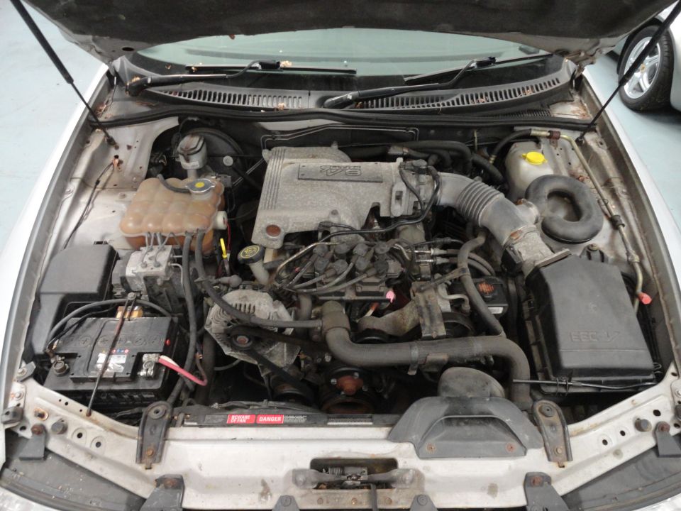 Car engine bay with hood open. Shows engine, battery, hoses, and air intake