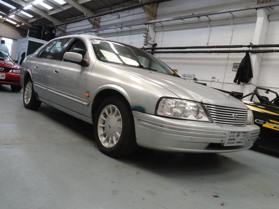 Silver sedan with chrome grille and multi-spoke wheels parked indoors. Another car is visible to the left