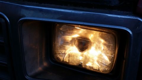 1981 Ford Bronco - A close-up view of a lit headlight on a vehicle, showing the textured glass and chrome trim surrounding it.
