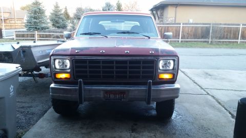 1981 Ford Bronco - The front view of a red Ford Bronco with its headlights and amber turn signals illuminated. A boat on a trailer is visible to the left of t…