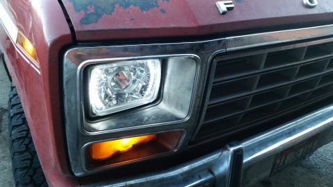 1981 Ford Bronco - Close-up of the front right headlight and amber turn signal of a red Ford truck. The vehicle has a chrome grille and bumper, and the hood s…