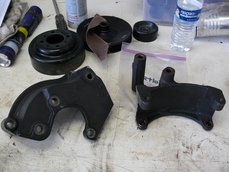 Two black metal automotive brackets on a workbench with tools and a water bottle.