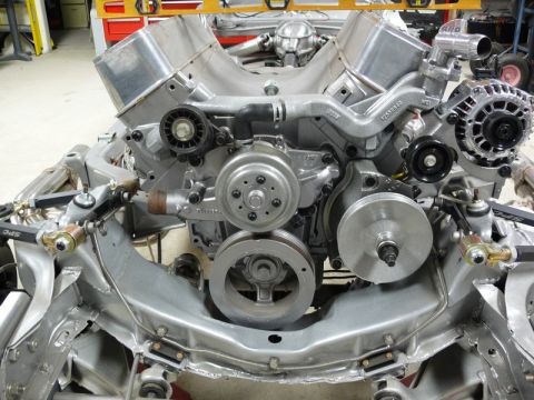 Silver V8 engine assembly in a vehicle frame with pulleys, alternator, and headers.