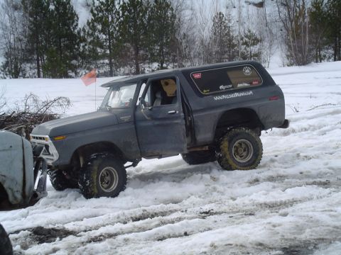 1993 Ford Bronco - A dark gray Ford Bronco navigates a snowy terrain with its driver's door ajar. Another vehicle is partially visible to the left.