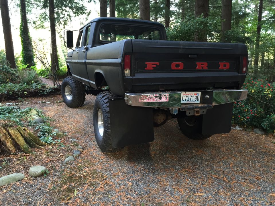 Lifted black 1970s Ford F-250 pickup truck with red "FORD" tailgate lettering, parked in a wooded area.