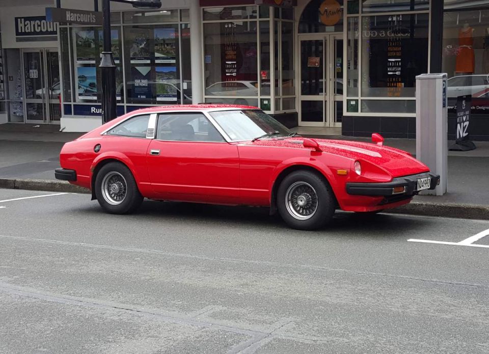 Red vintage coupe with fender mirrors and aftermarket wheels parked on a wet street.