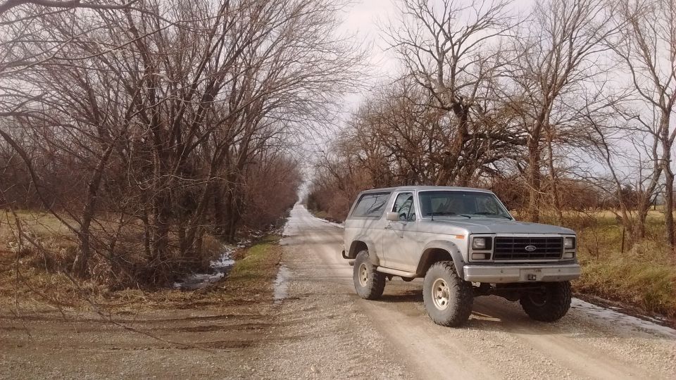 Silver 1980s Ford Bronco on a gravel road with bare trees.