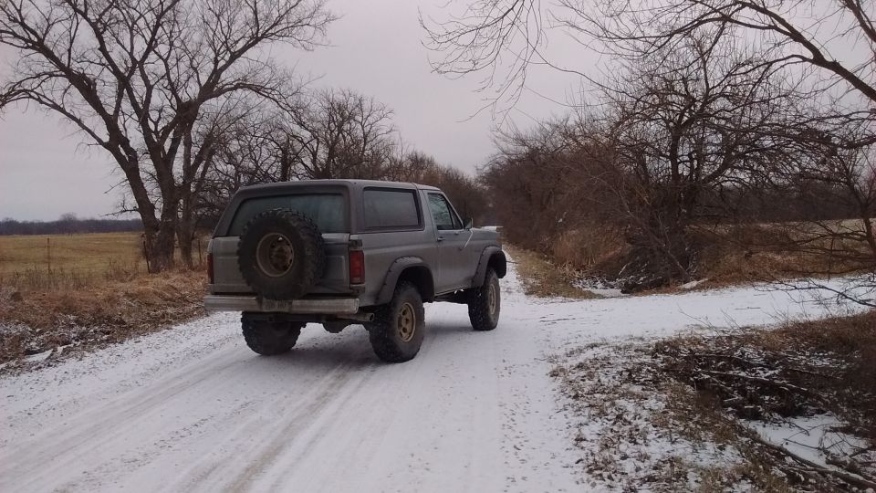 Gray 1985 Ford Bronco on a snowy road with bare trees.