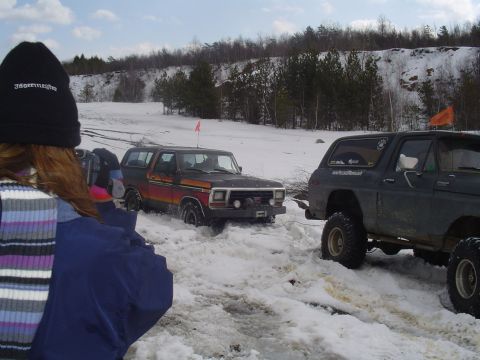 1993 Ford Bronco - Two SUVs navigate a snowy terrain with trees and a rocky hill in the background.