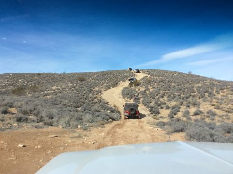 1979 Ford Bronco - A line of off-road vehicles, including a red Jeep Wrangler in the foreground, navigate a dusty dirt trail up a barren, scrub-covered hill u…