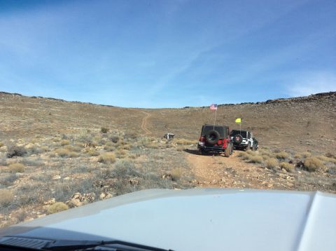 1979 Ford Bronco - Three Jeep Wranglers drive off-road on a dirt trail with sparse vegetation and a clear blue sky. The lead Jeep is red and has an American f…