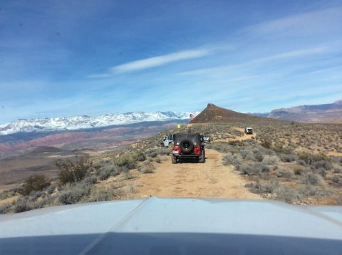 1979 Ford Bronco - A red Jeep Wrangler with an American flag drives on a dirt road in a desert landscape with snow-capped mountains in the background. The pho…