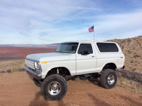 1979 Ford Bronco - A lifted white 1979 Ford Bronco is parked on a dirt hill with an American flag flying from a pole on its roof. The vehicle is viewed from a…