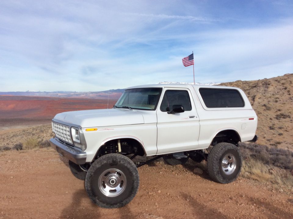 Lifted white 1979 Ford Bronco with American flag on a dirt hill.