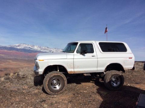 1979 Ford Bronco - A white, lifted 1979 Ford Bronco is parked on a dirt hill overlooking a valley with snow-capped mountains in the distance. An American flag…