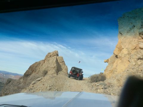 1979 Ford Bronco - A red Jeep Wrangler is driving up a rocky, off-road trail with a large rock formation to the left and a steep cliff face to the right. The…