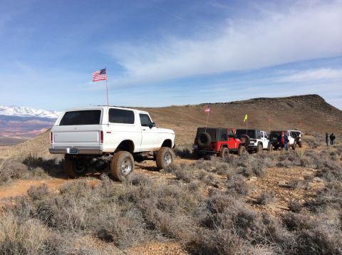 1979 Ford Bronco - A lifted white 1979 Ford Bronco is parked on a dirt trail in a desert landscape, with several Jeeps following behind it. An American flag i…