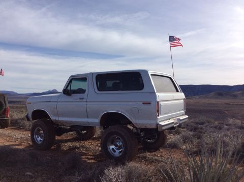 1979 Ford Bronco - A lifted, silver first-generation Ford Bronco is parked in a dry, desert landscape with an American flag flying from a pole behind it.