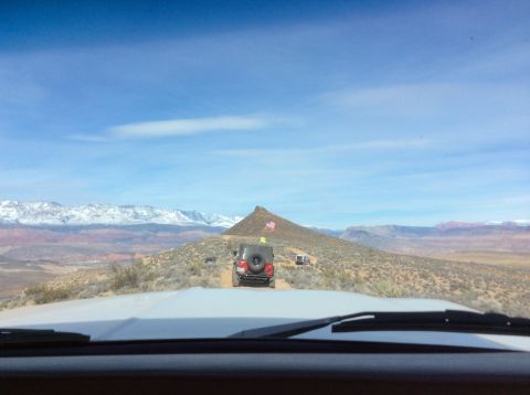 1979 Ford Bronco - A group of off-road vehicles, including a red Jeep Wrangler with American flags, drive along a dusty trail in a desert landscape with snow-…
