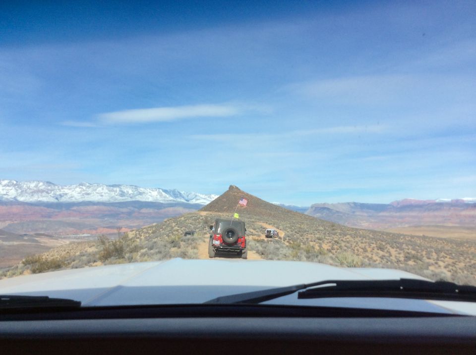 Red Jeep Wrangler with American flags driving on a desert trail with mountains in the background.