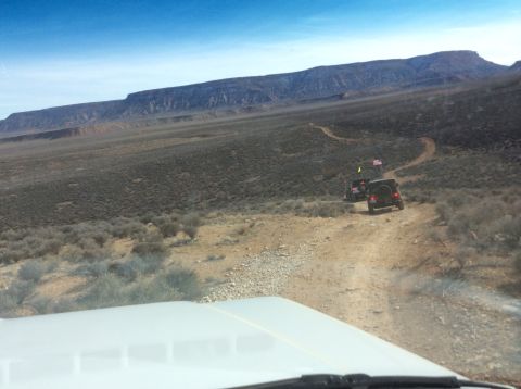 1979 Ford Bronco - Two off-road vehicles with flags drive up a dusty, winding trail in a barren, mountainous landscape under a clear blue sky. The front of a…