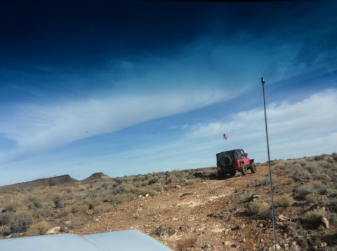 1979 Ford Bronco - A red Jeep Wrangler is driving on a dirt road in a desert landscape under a bright blue sky with wispy clouds. An American flag is attached…