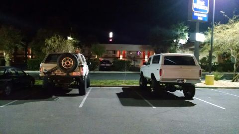1979 Ford Bronco - Two lifted SUVs are parked in a lot at night in front of a motel. The SUV on the left is a light color with a spare tire mounted on the rea…