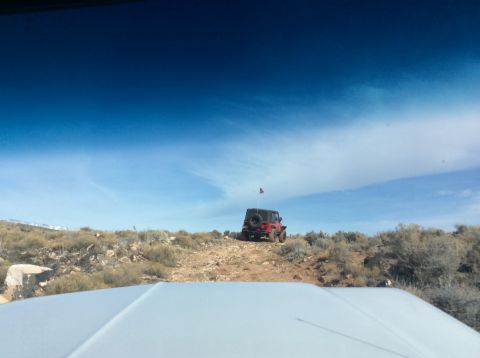 1979 Ford Bronco - A red Jeep Wrangler is parked on a rocky, arid hillside under a bright blue sky with scattered clouds. The vehicle has an American flag mou…