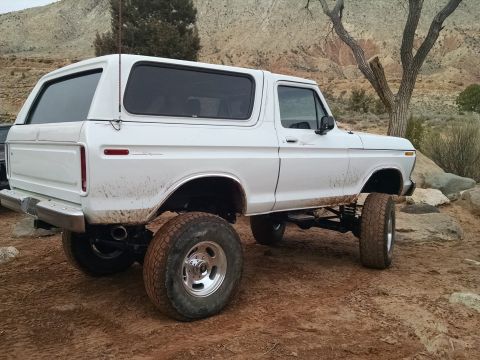 1979 Ford Bronco - A white, lifted 1979 Ford Bronco is parked on a dirt road with rocky hills and sparse vegetation in the background. The vehicle is covered…