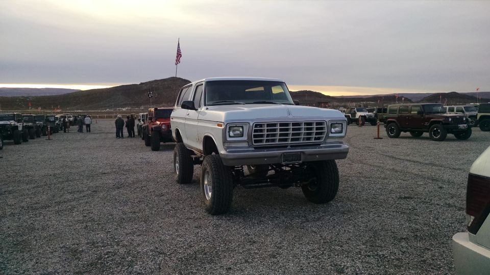 Lifted white 1979 Ford Bronco parked in gravel lot with other vehicles and American flag.