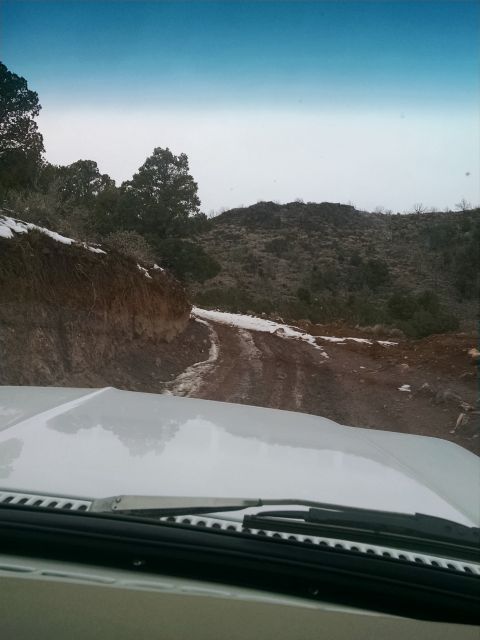 1979 Ford Bronco - View from the driver's seat of a white vehicle driving on a muddy and snowy dirt road through a hilly, wooded area.