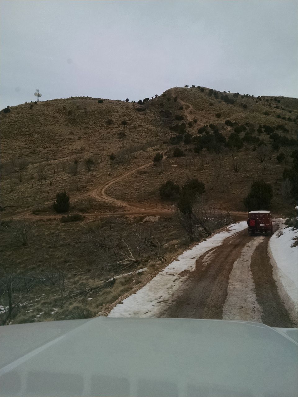 Red vintage SUV on a snowy dirt road ascending a dry, grassy hill with a distant communication tower.