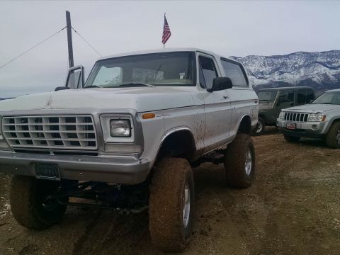 1979 Ford Bronco - A lifted, white 1979 Ford Bronco with large mud tires sits parked on a dirt lot with other vehicles and snow-capped mountains in the backgr…
